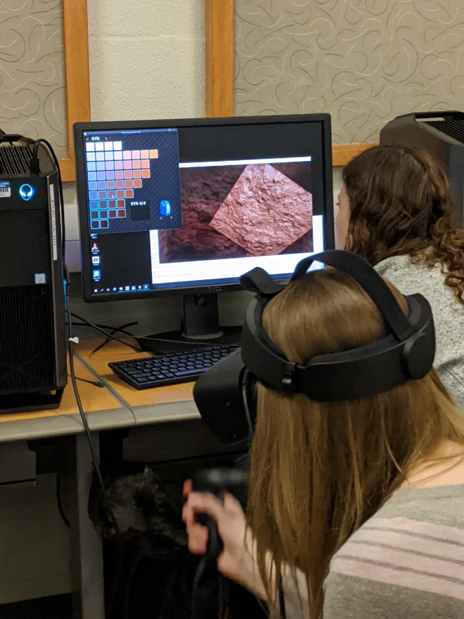 A participant wearing a VR headset faces a monitor showing a virtual excavation.