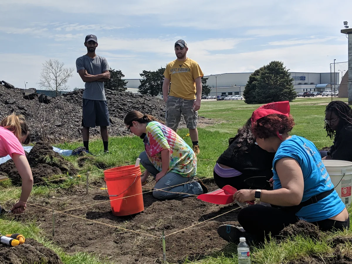 A group works beside marked excavation areas outdoors, with buckets and field tools.